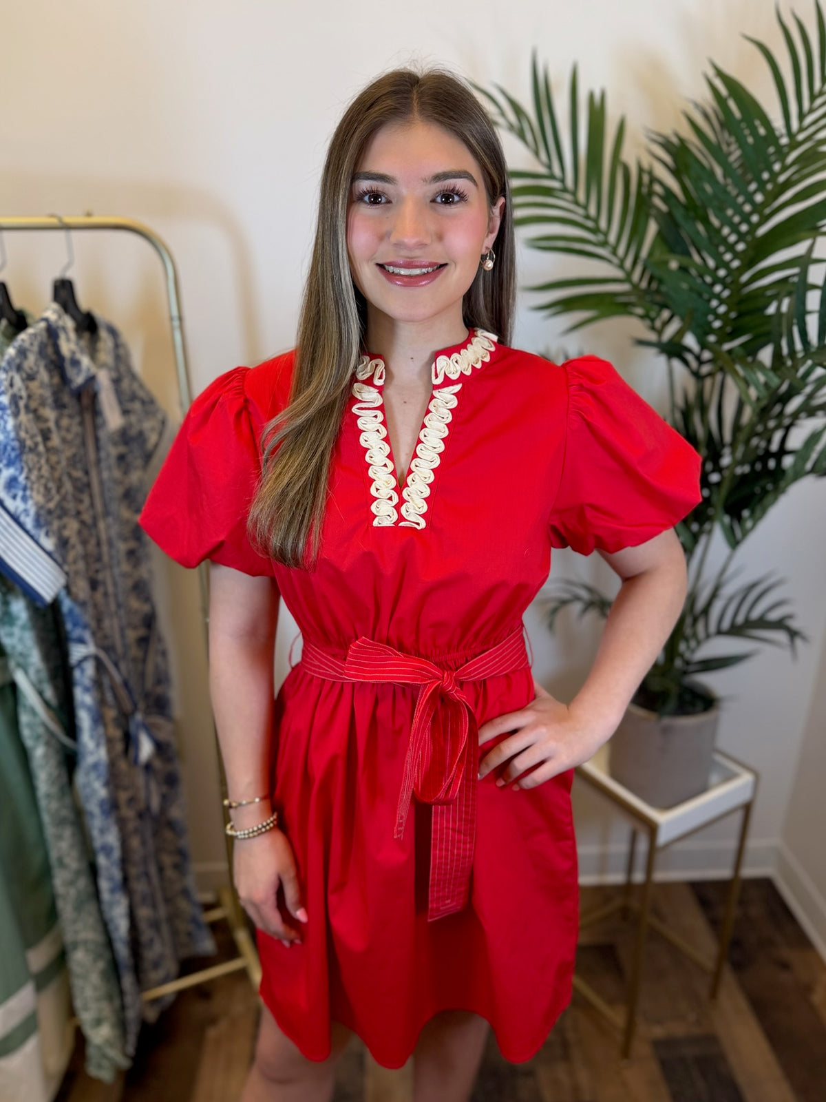 Woman wearing Red puff sleeve dress with self tie waist accent standing in a room with a plant and clothing rack.
