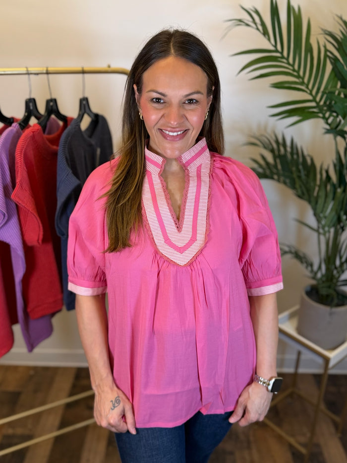 A woman wearing a bright pink short-sleeve blouse with a striped split V-neck trim, standing in a boutique setting with clothing racks and a potted plant in the background.