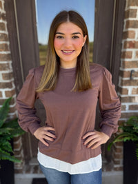 A woman wearing a mocha long-sleeve top with a white layered hem, standing in front of a brick door.