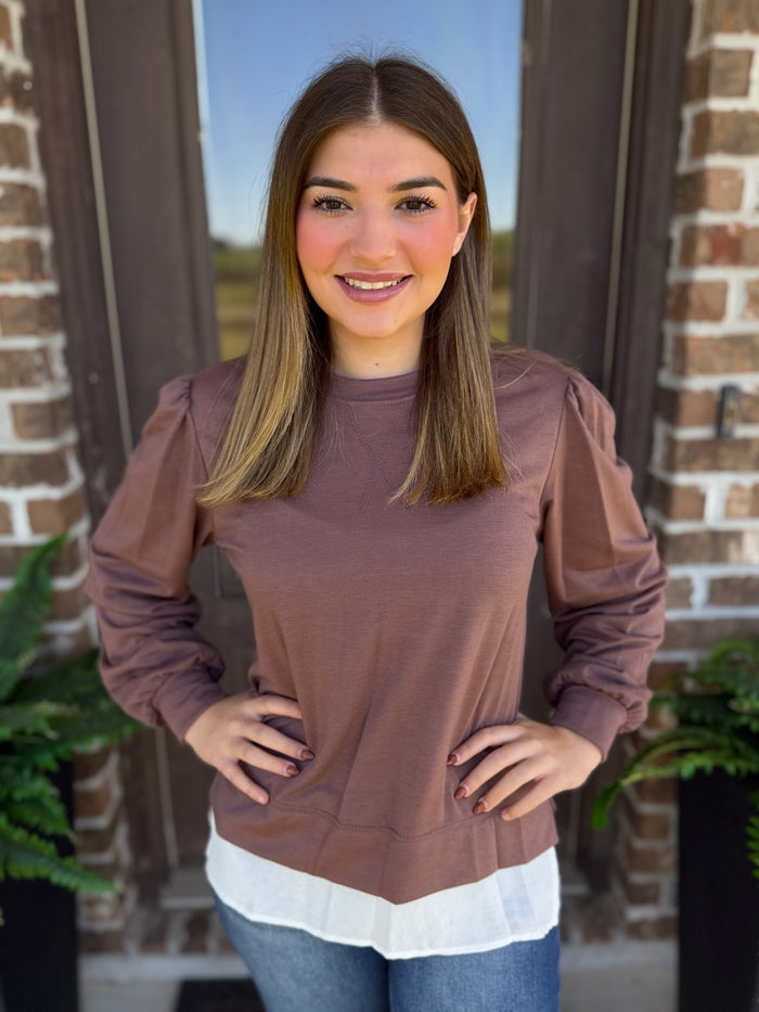A woman wearing a mocha long-sleeve top with a white layered hem, standing in front of a brick door.