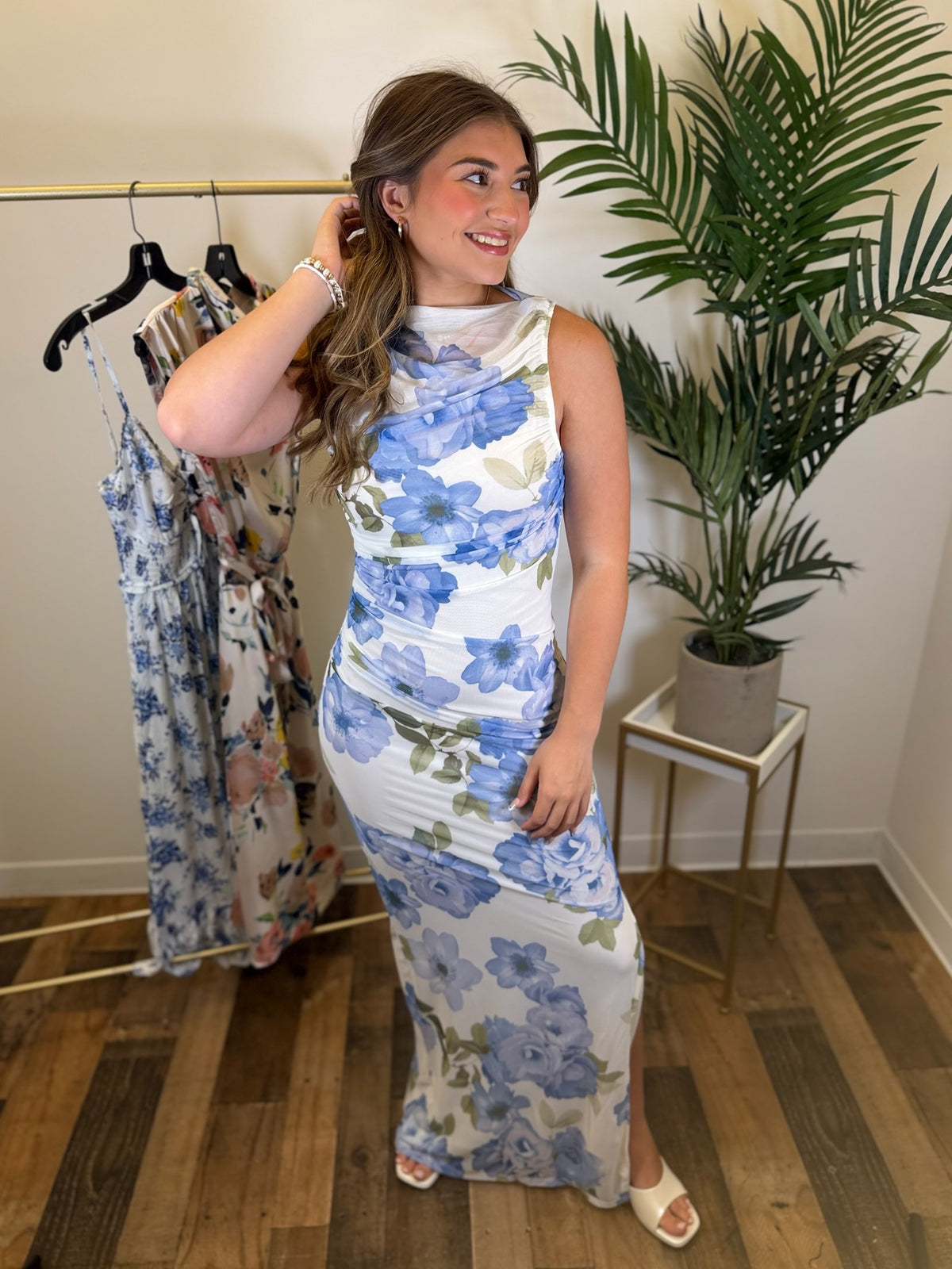 Woman wearing a sleeveless floral maxi dress standing in a room with a plant and rack of dresses.