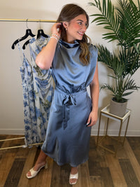 Woman wearing a slate blue midi dress with draped detailing standing in a room with a plant and clothing rack.