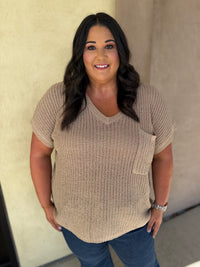 A woman wearing a beige short-sleeve knit top with a chest pocket, paired with blue jeans, standing against a stucco wall.