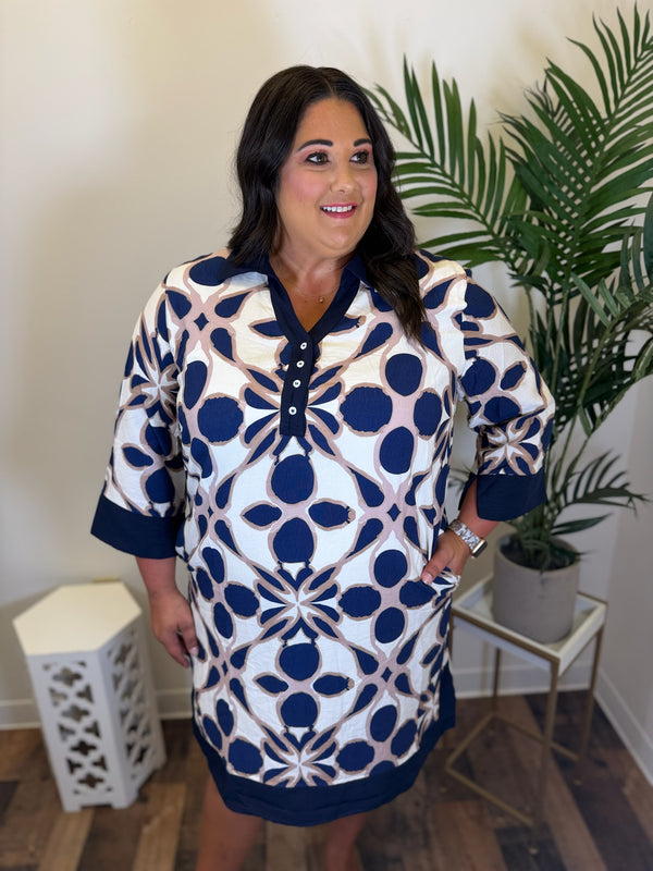A woman modeling a short-sleeve tunic dress with a navy blue and white geometric medallion pattern, standing in a room with a potted palm plant.