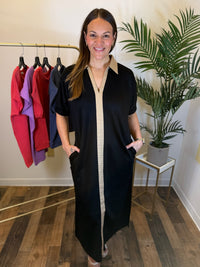 A woman modeling a black midi dress with a beige center panel and collar, standing in a boutique setting with a clothing rack and a potted plant in the background.