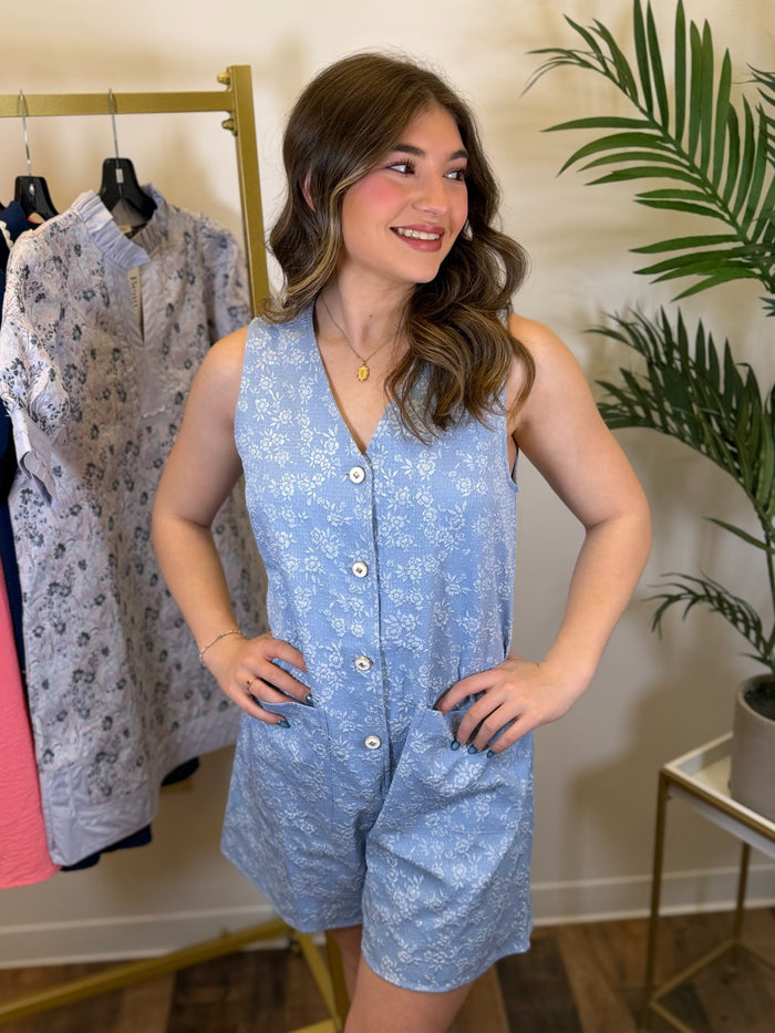 Woman wearing a sleeveless blue floral button front romper in a clothing store setting.