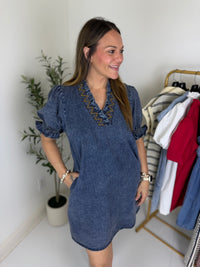 Woman wearing a dark blue denim mini dress with embroidery in a room with a clothing rack and plant.