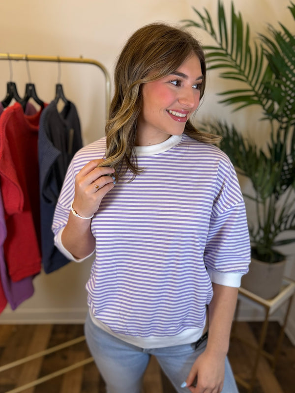 A woman wearing a lavender and white horizontally striped crewneck short sleeve top, standing in a boutique setting with a clothing rack and a potted plant in the background.