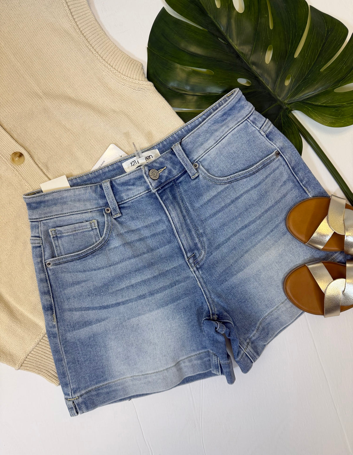 High rise, light wash denim shorts on a white surface with a beige cardigan and gold sandals.