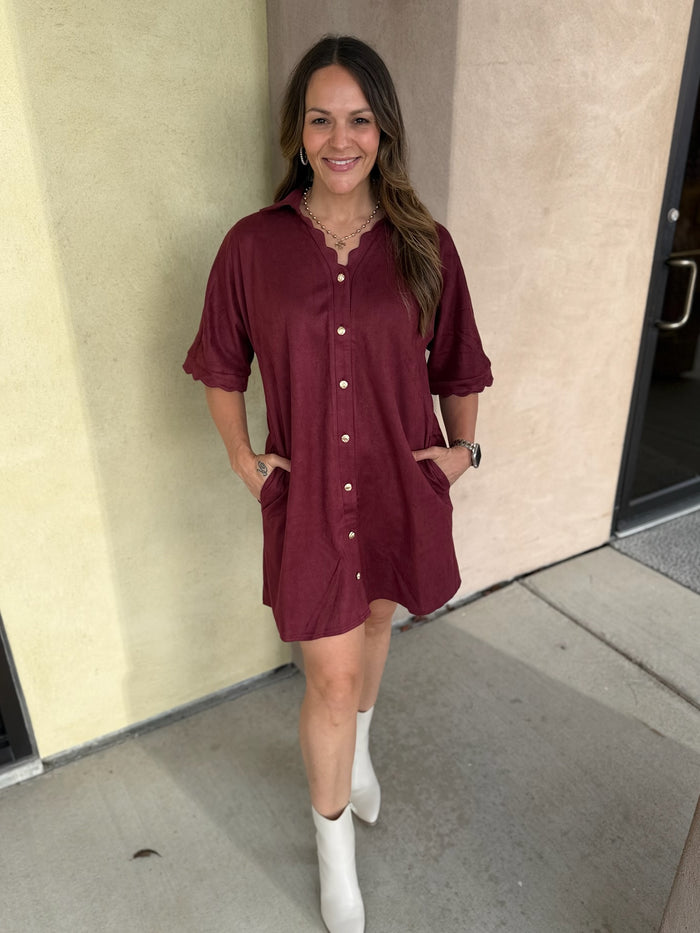 A woman wearing a short-sleeve burgundy button-front shirt dress with scalloped trim and pockets, paired with white ankle boots, standing outdoors against a beige stucco wall.