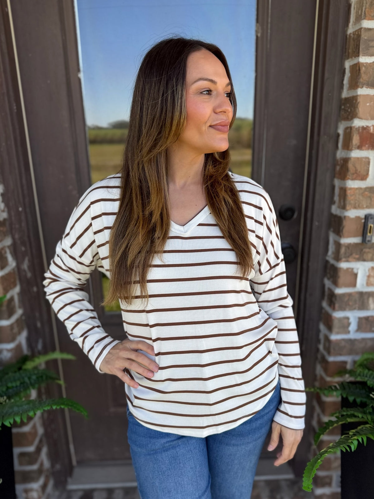 A woman wearing a long-sleeve V-neck top with thin brown horizontal stripes on an off-white base, paired with blue jeans, standing in front of a brick doorway.