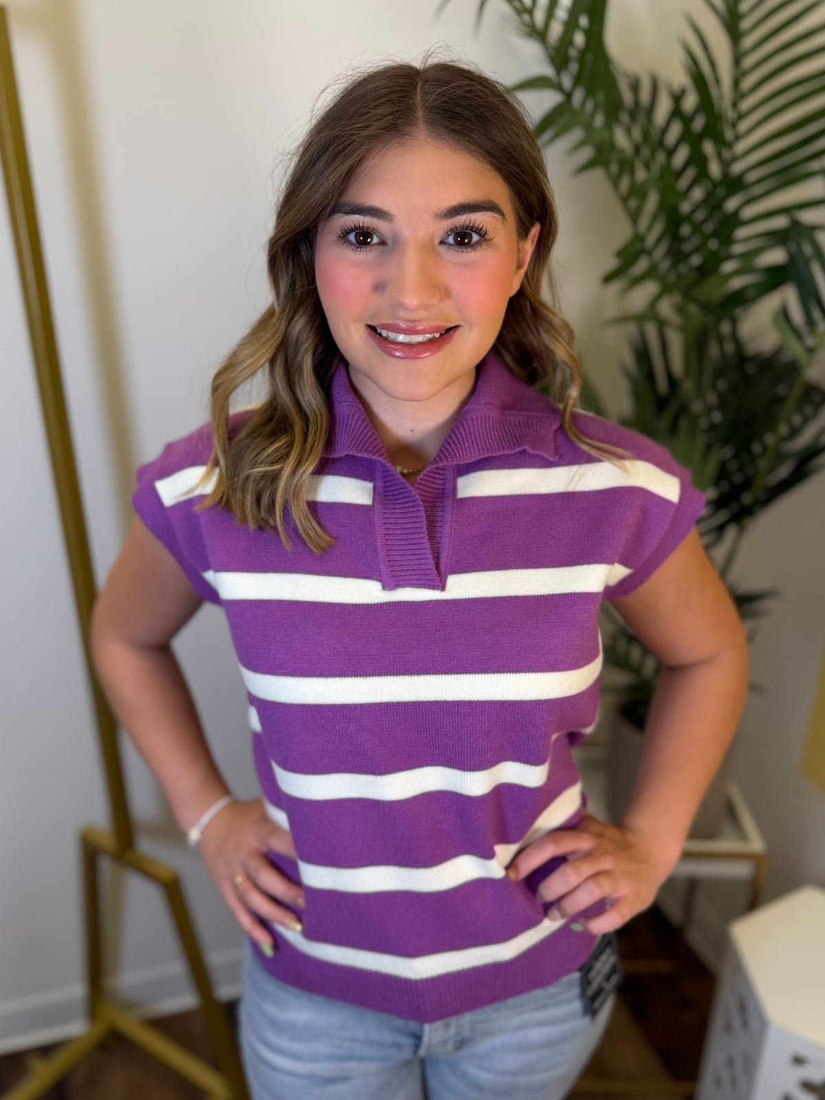 A smiling young woman wearing a purple and white horizontally striped short-sleeve polo shirt, paired with light blue jeans, standing in an indoor setting with a plant and clothing rack in the background.