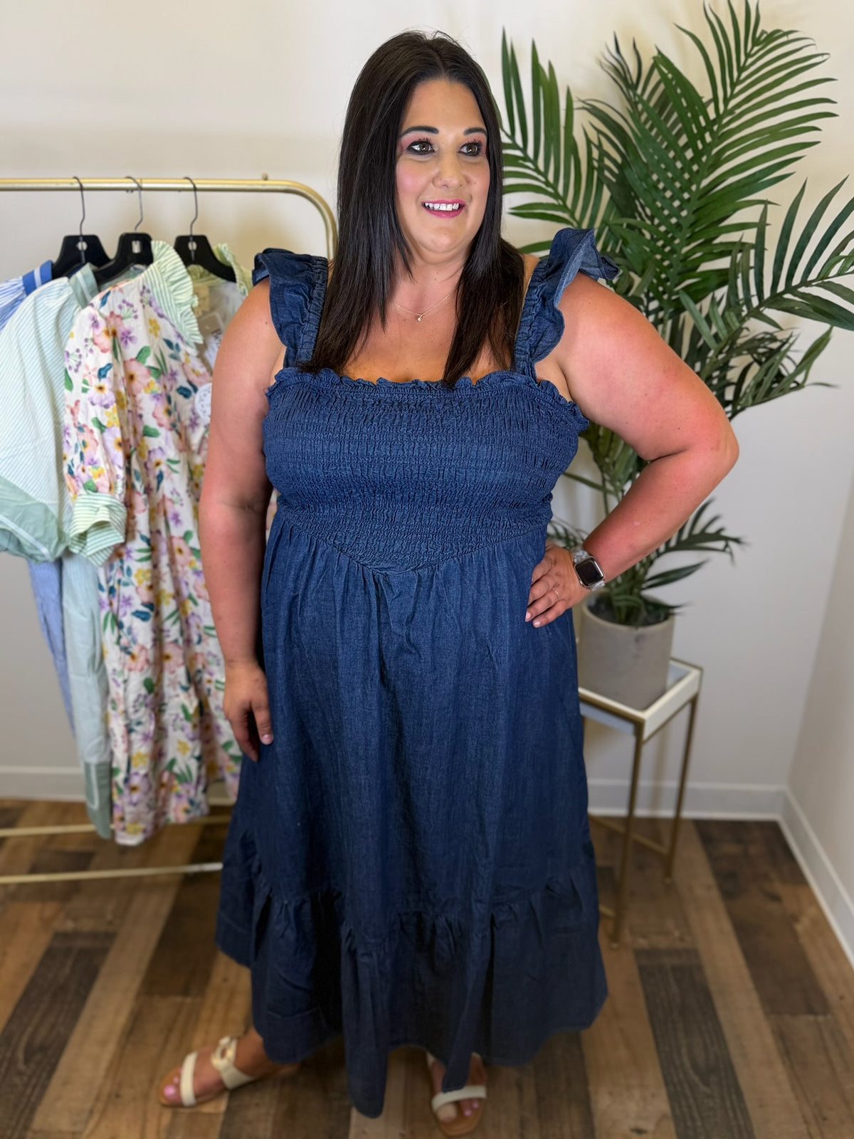 Woman wearing a blue denim maxi dress with ruffle straps and smocked bodice standing in a room with a clothing rack and plants.