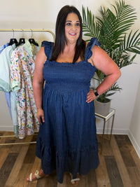 Woman wearing a blue denim maxi dress with ruffle straps and smocked bodice standing in a room with a clothing rack and plants.
