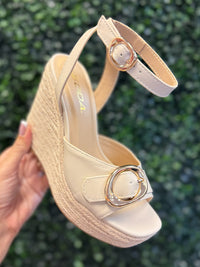 Beige sandal with decorative buckle held against a green leafy background