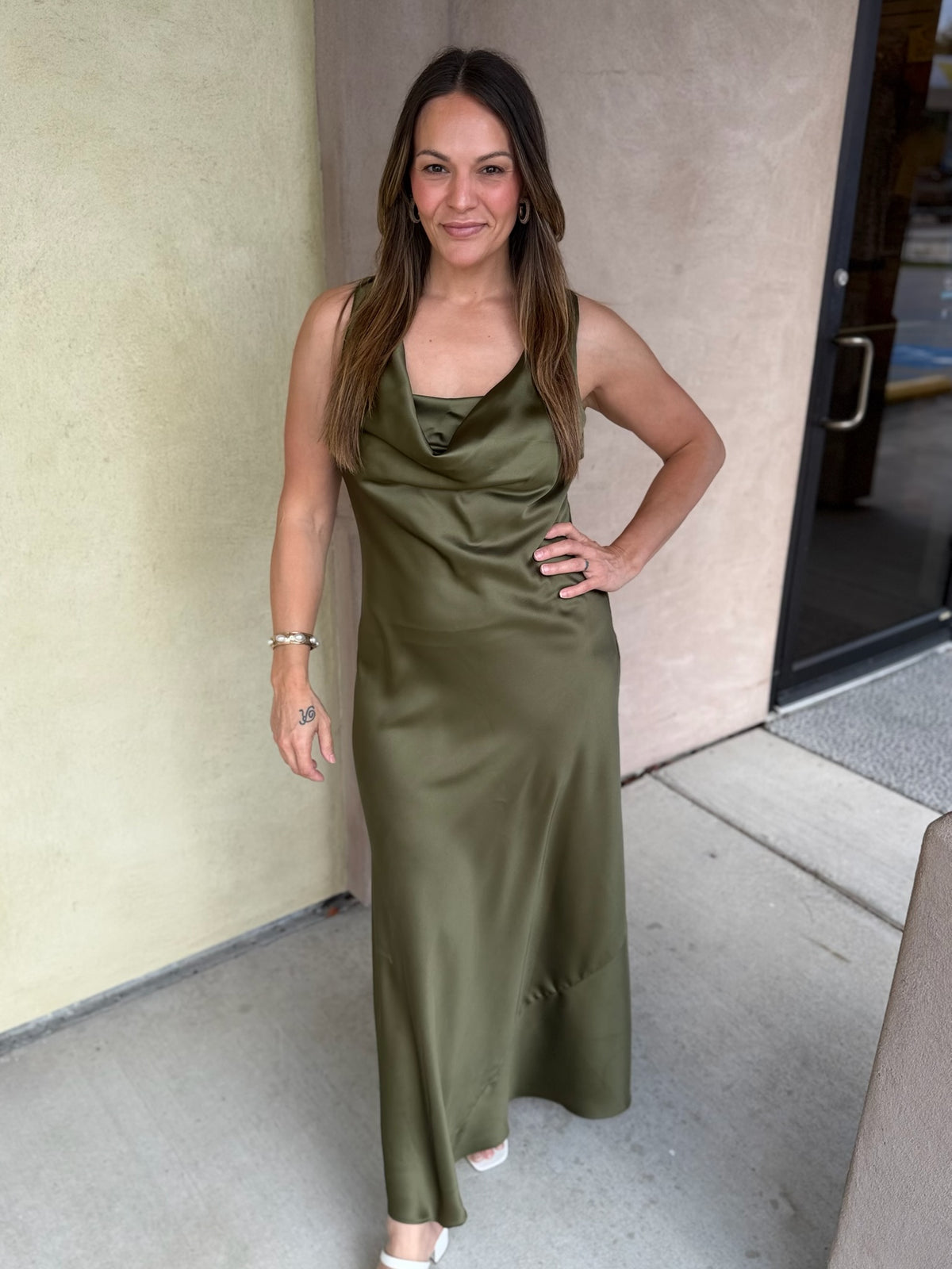 Olive green satin midi dress with spaghetti straps and draped neckline worn by a woman standing in front of beige building.