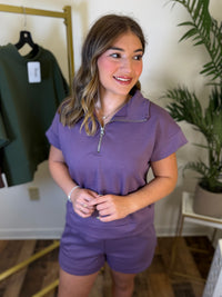 A woman wearing a matching purple short-sleeve quarter-zip pullover and shorts set, standing in a boutique setting with a clothing rack and potted plants in the background.