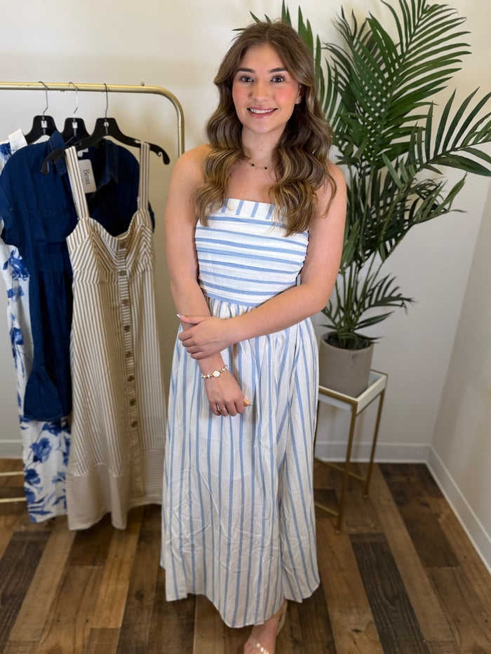 Woman in a strapless blue and white striped midi dress standing in a clothing store with racks of dresses and plants.
