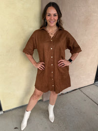 A woman wearing a short-sleeve camel button-front shirt dress with scalloped trim and pockets, paired with white ankle boots, standing outdoors against a beige stucco wall.