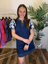 A woman modeling a navy blue mini dress with white leaf-patterned short sleeves and thin white horizontal stripes near the hem, standing in a boutique setting with a clothing rack and potted plant in the background.
