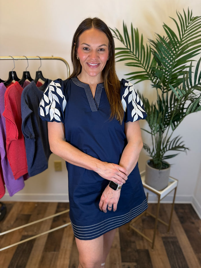 A woman modeling a navy blue mini dress with white leaf-patterned short sleeves and thin white horizontal stripes near the hem, standing in a boutique setting with a clothing rack and potted plant in the background.