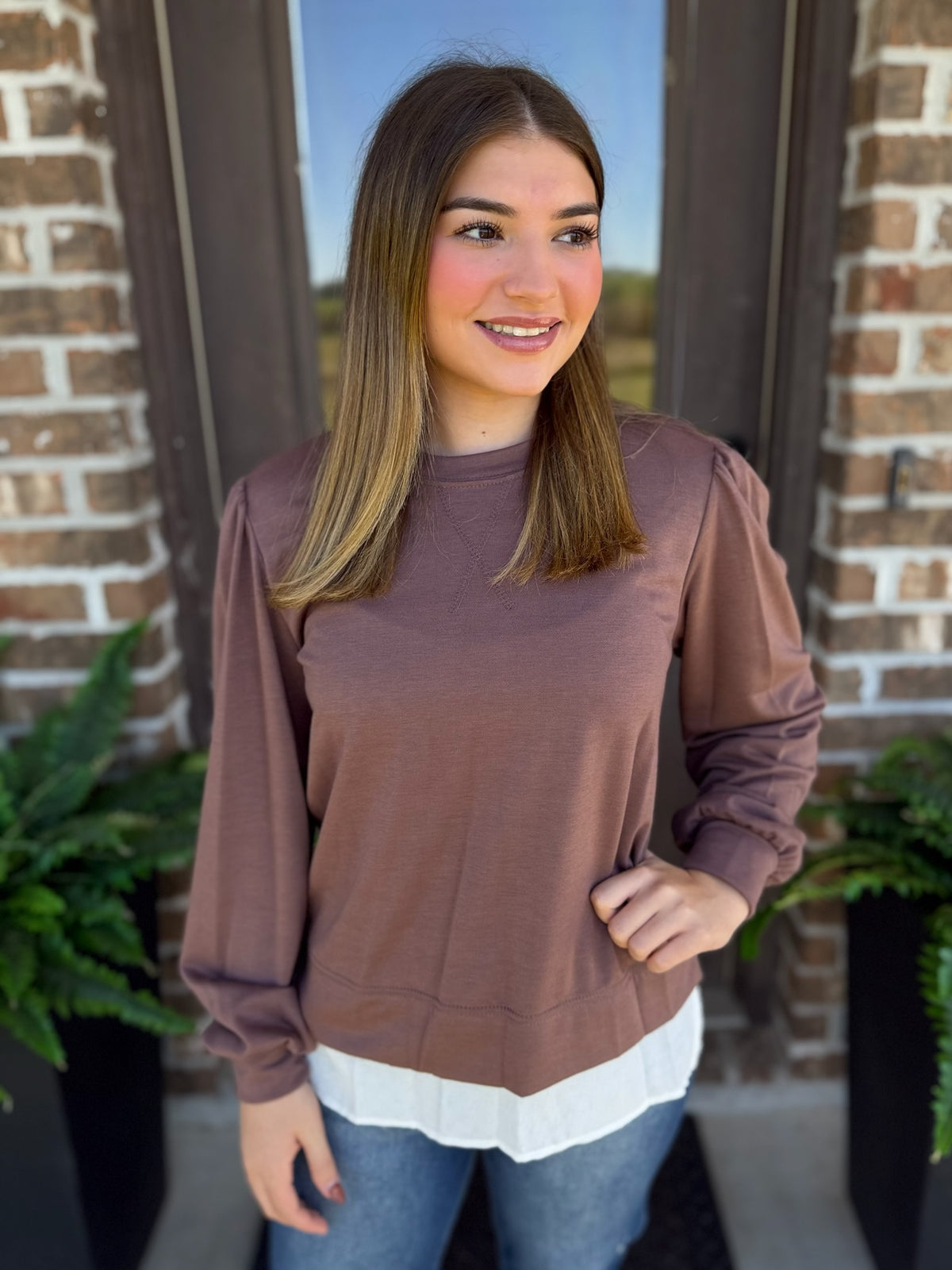 A woman wearing a mocha long-sleeve crewneck sweatshirt over a white top and blue jeans, standing in front of a brick doorway.