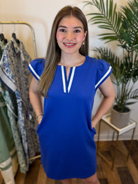 Woman wearing a royal blue v neck dress with ruffled cap sleeves standing in a room with a clothing rack and plant.
