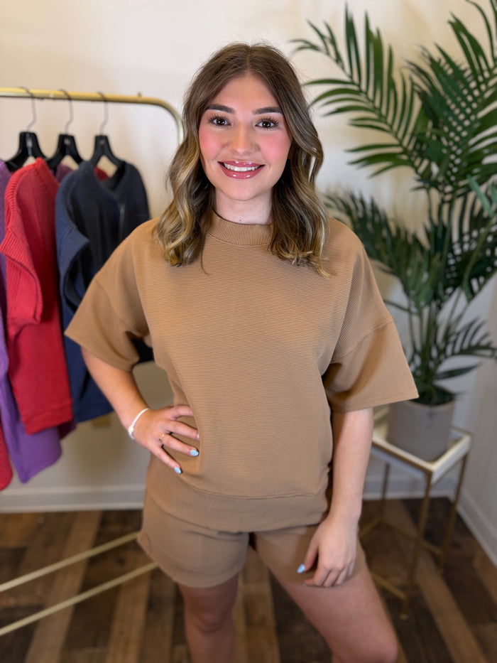 A woman modeling a matching tan short-sleeve top and shorts set in an indoor boutique setting with a clothing rack and a potted plant in the background.