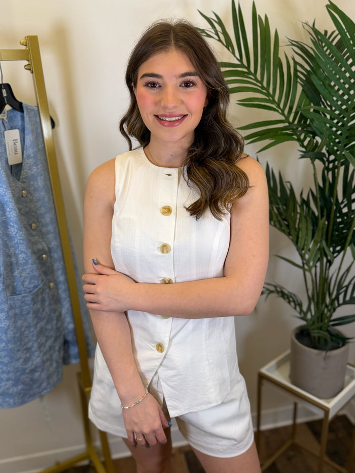 Woman in a white outfit standing in a room with a plant and clothing rack.