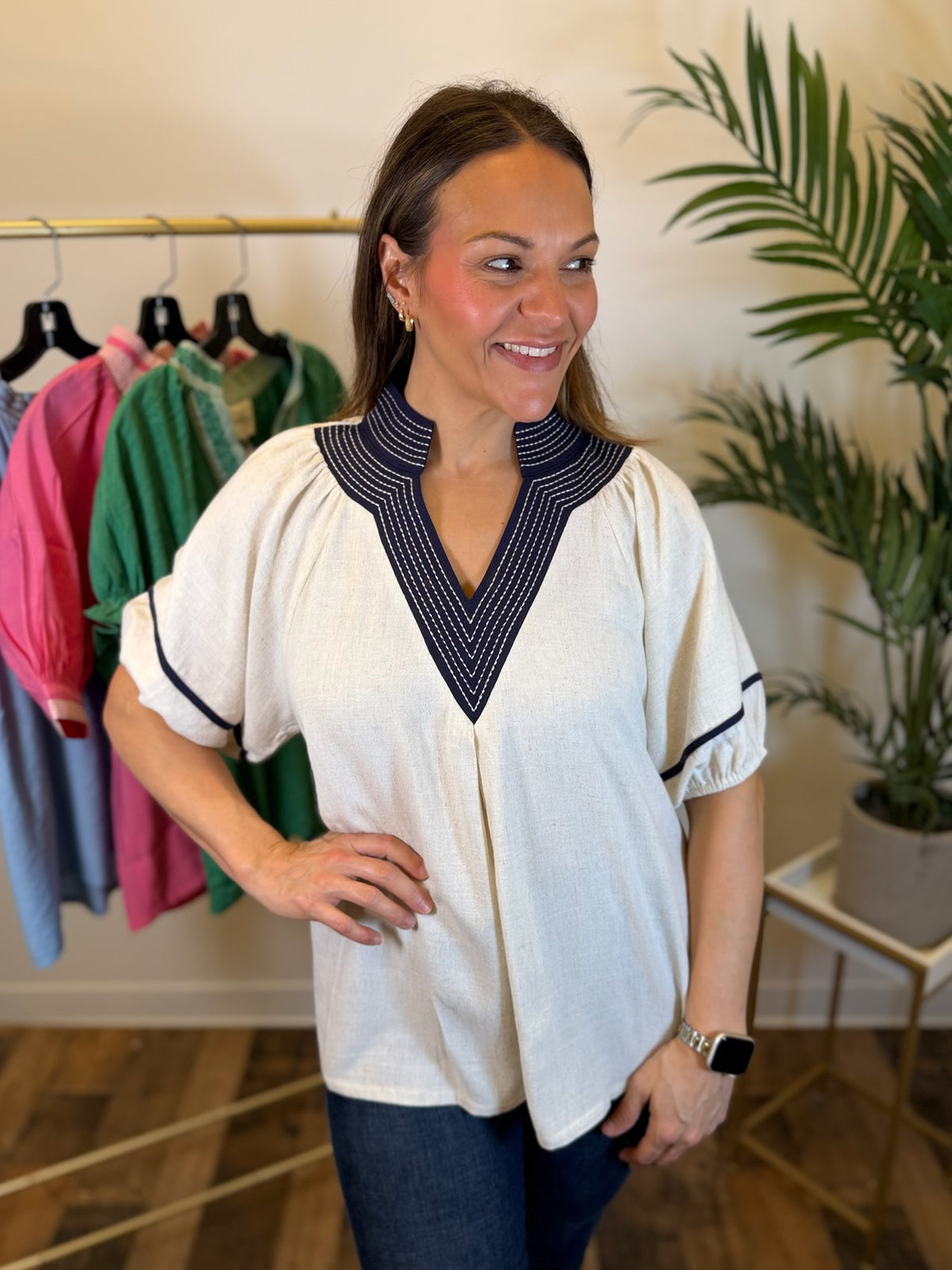 A woman modeling a cream-colored short-sleeve blouse with a navy blue and white patterned V-neck collar and sleeve trim, paired with dark jeans in a boutique setting.