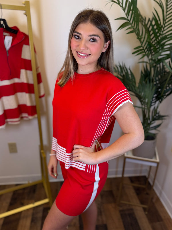 A woman modeling a bright red short-sleeve dress with white striped accents on the sleeves and hem, standing in an indoor boutique setting with a clothing rack and plant in the background.