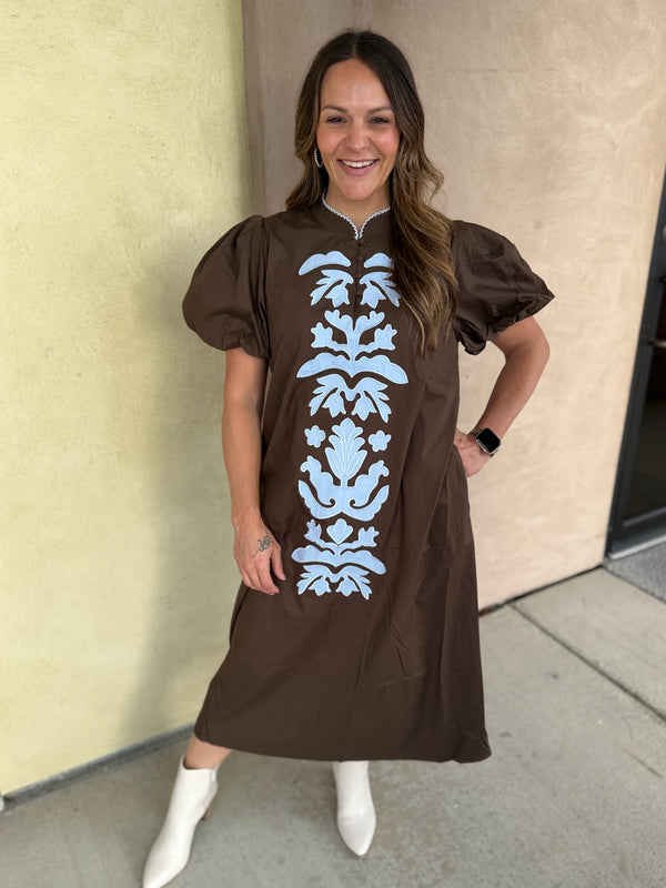 A woman wearing a brown midi dress with puff sleeves and a vertical light blue damask pattern on the front, paired with white ankle boots, standing outdoors against a beige wall.