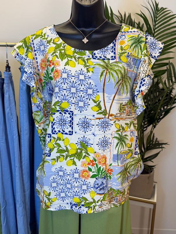 Colorful floral and tropical patterned cap sleeve top in shades of Blue, Yellow, Green, and orange on a hanger with a plant in the background