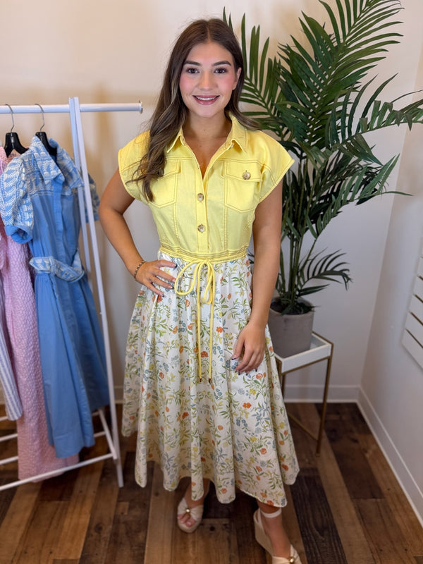 Woman wearing a midi dress with yellow top and floral skirt in a room with clothing racks and plants.