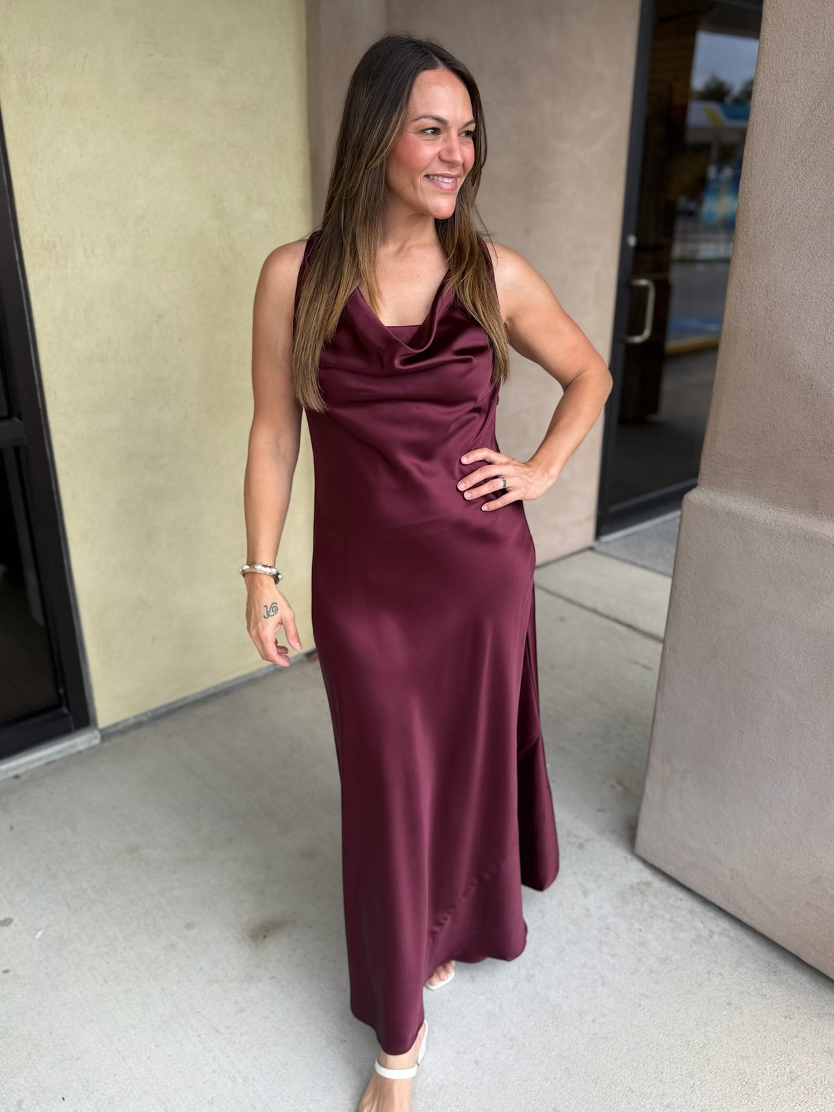 Burgundy satin midi dress with spaghetti straps and draped neckline worn by a woman standing in front of beige building.