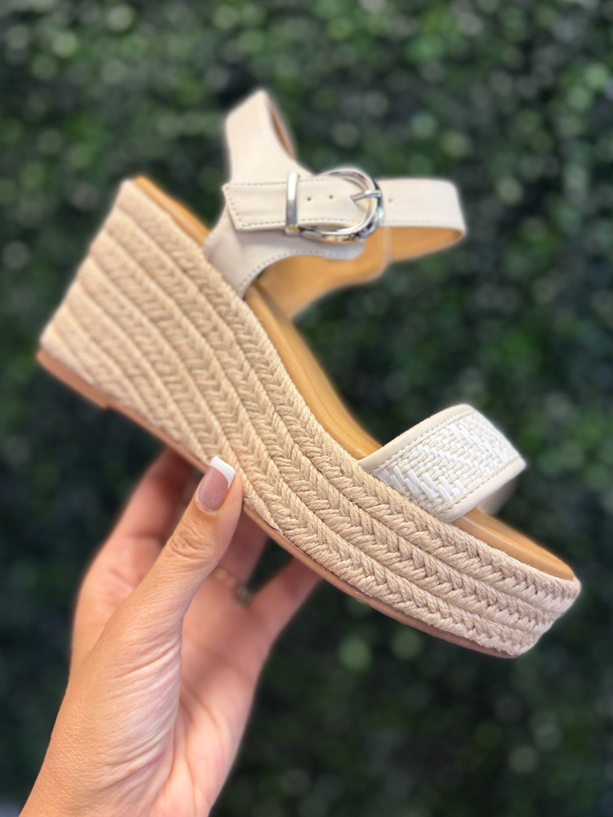 Beige wedge sandal with espadrille sole held against a blurred green background