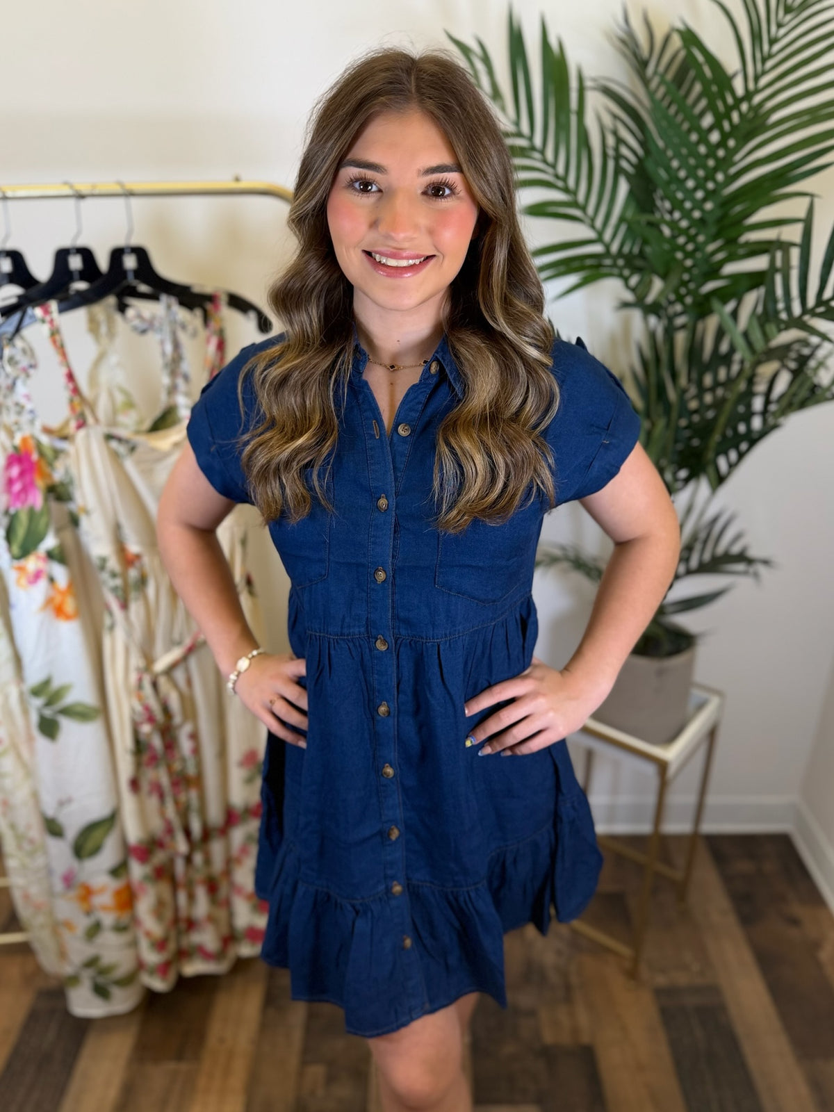 Woman wearing a blue chambray button front dress with babydoll design and short sleeves in a clothing store with plants in the background