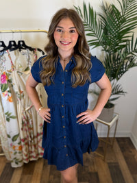 Woman wearing a blue chambray button front dress with babydoll design and short sleeves in a clothing store with plants in the background