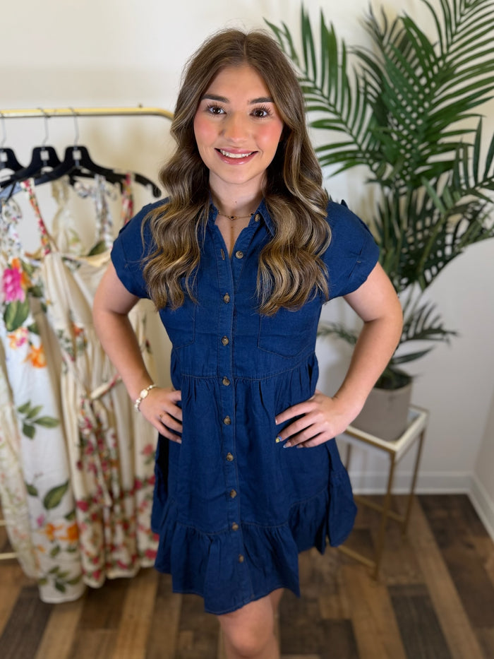 Woman wearing a blue chambray button front dress with babydoll design and short sleeves in a clothing store with plants in the background