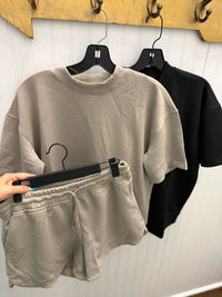 A taupe/beige crewneck t-shirt and matching shorts set hanging on black hangers against a white paneled wall, with a hand holding up a matching pair of shorts in the foreground. A black t-shirt is visible in the background.