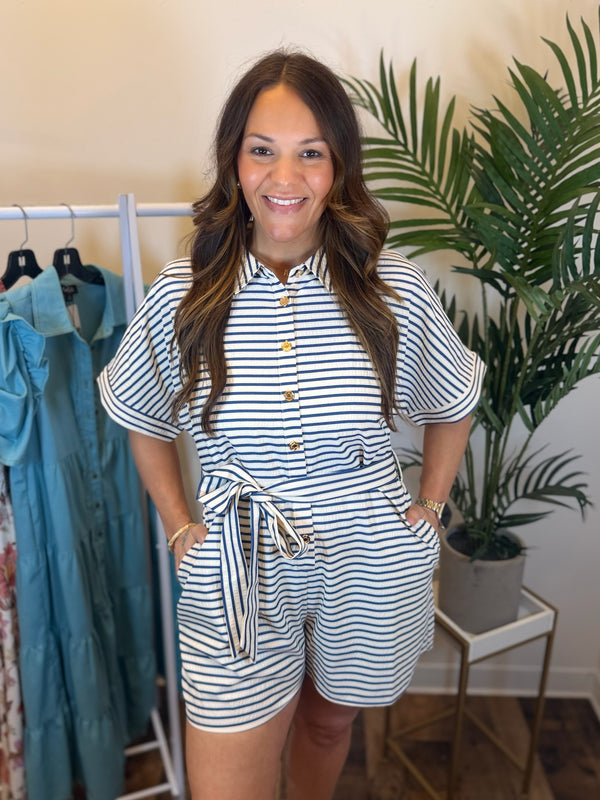 Woman wearing a striped romper with gold button details, collar neck, and matching fabric belt standing in a room with clothing and plants.