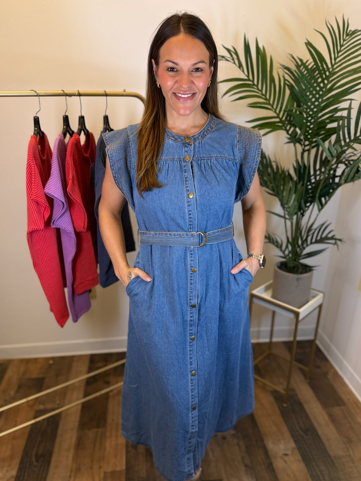 A woman modeling a blue denim midi dress with a button-down front, ruffled shoulders, and a matching belt, standing in a boutique setting with a clothing rack and a potted plant in the background.