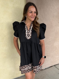A woman wearing a black short-sleeve dress with leopard print trim on the neckline and hem, standing against a beige stucco wall.