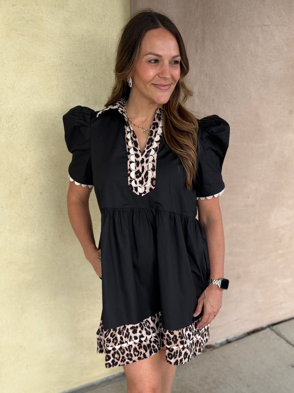 A woman wearing a black short-sleeve dress with leopard print trim on the neckline and hem, standing against a beige stucco wall.