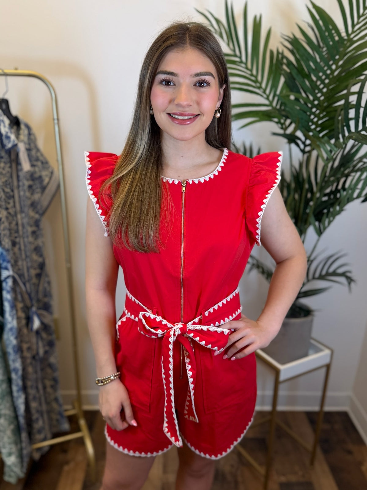 Woman wearing a red cap sleeve romper with white trim in a room with plants and a mirror.