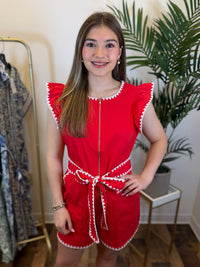 Woman wearing a red cap sleeve romper with white trim in a room with plants and a mirror.