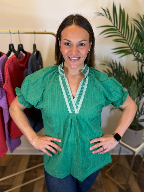 A woman wearing a green short-sleeve blouse with embroidered trim on the V-neck, standing in a boutique with clothing racks and a plant in the background.