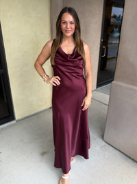 Burgundy satin midi dress with spaghetti straps and draped neckline worn by a woman standing in front of beige building.