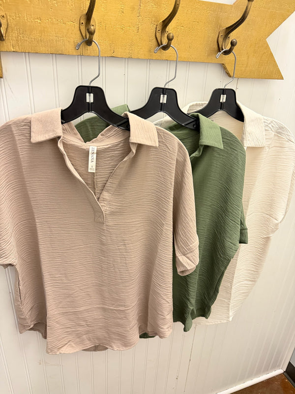 Three women's short-sleeve collared woven tops hanging on black hangers against a white paneled wall, with a yellow peg rack above. The front top is light beige, the middle is olive green, and the back is off-white/cream.