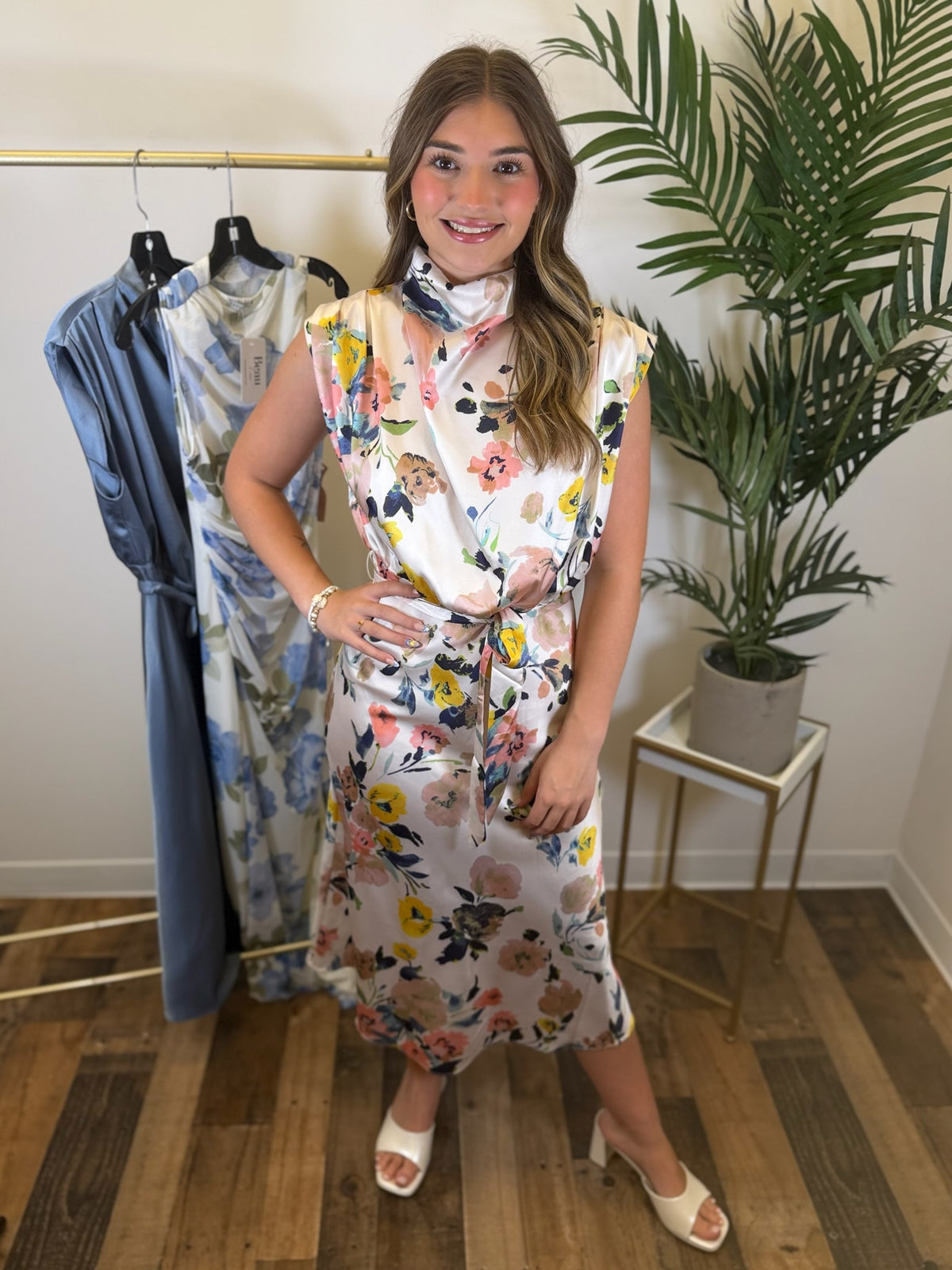 Woman wearing a floral midi dress with draped neckline standing in a room with clothing on a rack and a plant.